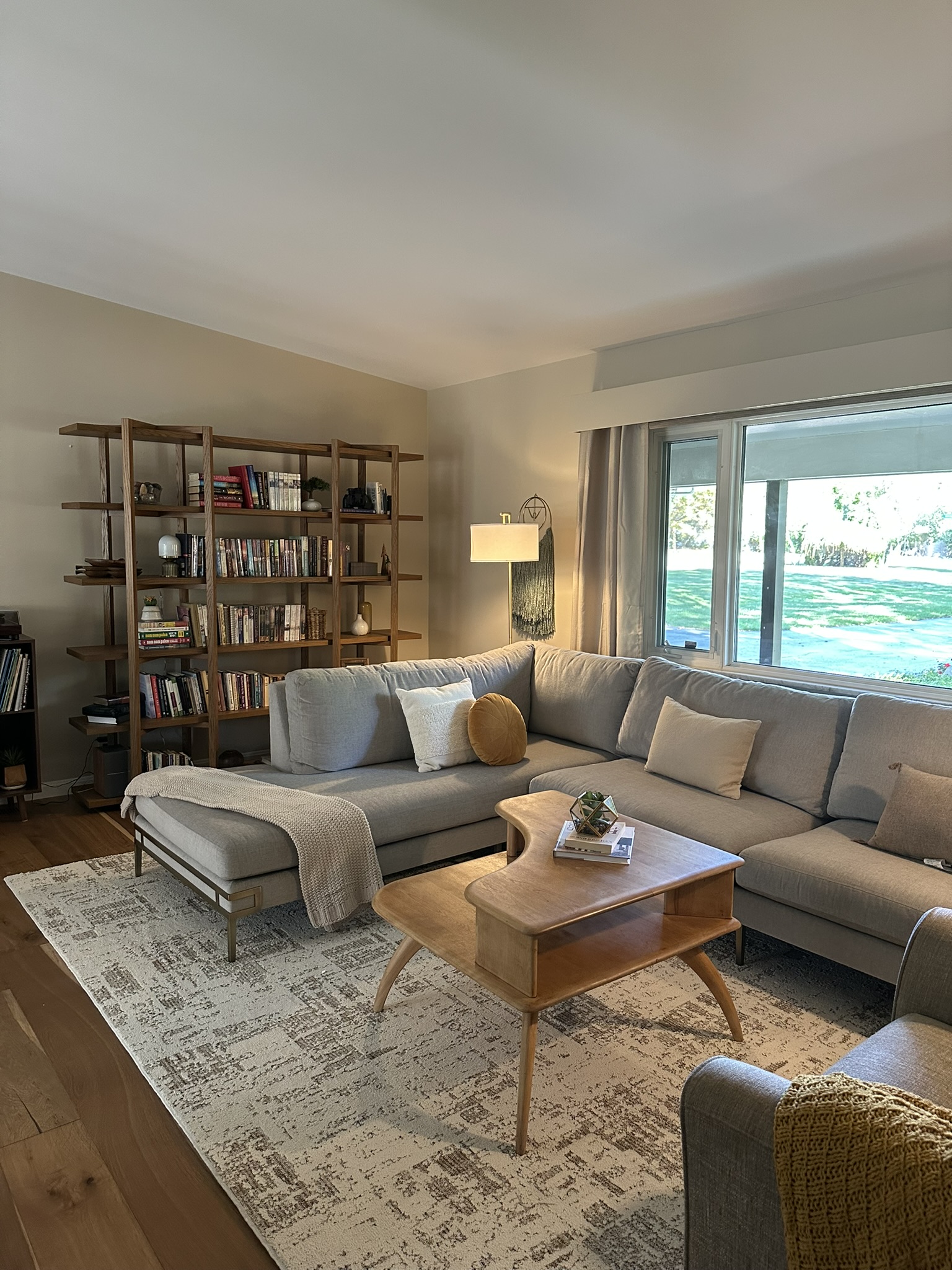 IMG_9617 A cozy living room with a gray sectional sofa, matching armchair, two floor lamps, a wooden coffee table, and large windows letting in natural light. A neutral custom area rug finished with true serging from Yoder Floors covers the wooden floor for added warmth.