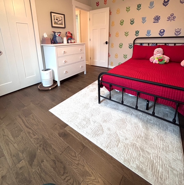 Children's bedroom decorated with floral wallpaper, iron twin bed with red linens, and a velvety soft white area rug over oak hardwood floors.